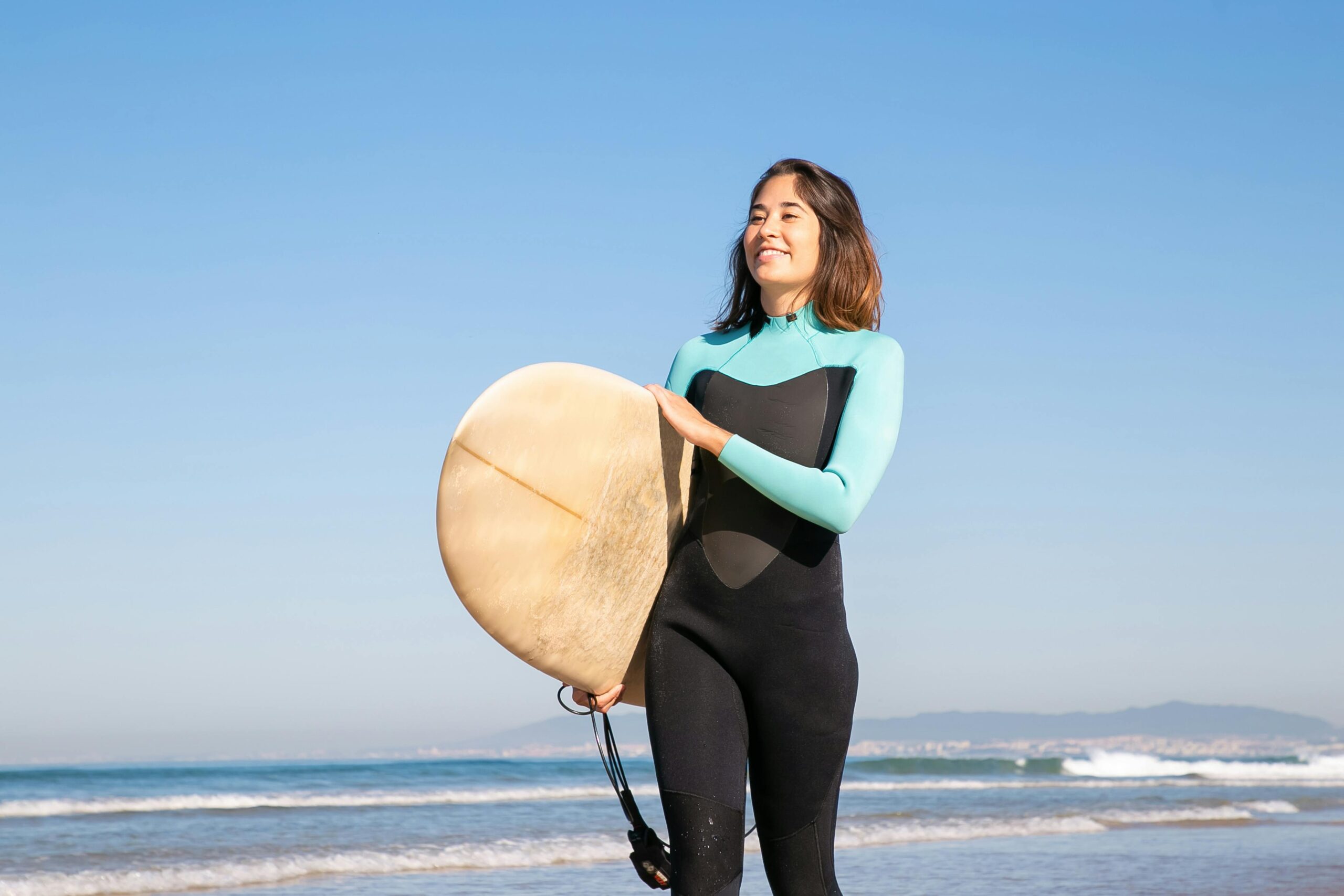A joyful woman in a wetsuit holding a surfboard on a sunny beach in Portugal.